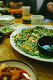 A plate of savory Korean pancakes garnished with green onions and served with a small dish of dipping sauce on the side. The table also features small side dishes of pickled vegetables, a cup, and various cups and plates in a casual dining setting.