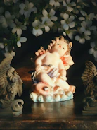 A small cherub-like figurine with rosy cheeks and a peaceful expression is surrounded by white flowers. The figure is sitting on a textured base, flanked by two decorative bird sculptures. The scene is warmly lit, highlighting the soft pastel colors of the figurine and creating a serene atmosphere.