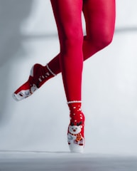 A pair of legs in red tights and festive socks featuring a reindeer design are posed en pointe. The background is a plain light color, highlighting the vibrant red and white of the attire.
