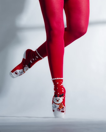 A pair of legs in red tights and festive socks featuring a reindeer design are posed en pointe. The background is a plain light color, highlighting the vibrant red and white of the attire.