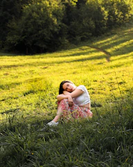 A thoughtful woman meditating outdoors surrounded by soft greenery and warm sunlight.