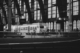 A black and white photograph of a train station with a train on the tracks. The station features large arched windows and numerous passengers on the platform, some walking, others waiting. Overhead signs indicate the platform numbers and destinations. The overall architecture is industrial with exposed beams and metal structures, contributing to an urban atmosphere.