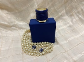 A dark blue, square-shaped bottle with a textured cap. Positioned among strands of pearl necklaces and two blue, star-shaped decorations. The background features a soft beige, textured fabric.