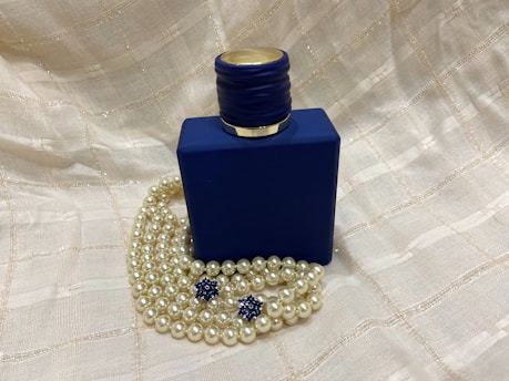 A dark blue, square-shaped bottle with a textured cap. Positioned among strands of pearl necklaces and two blue, star-shaped decorations. The background features a soft beige, textured fabric.