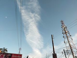 A tall telecommunications tower stands against a clear blue sky with a wisp of white cloud. Several power lines are visible, crisscrossing the scene. Part of an advertising billboard is visible in the corner with text in a foreign script.