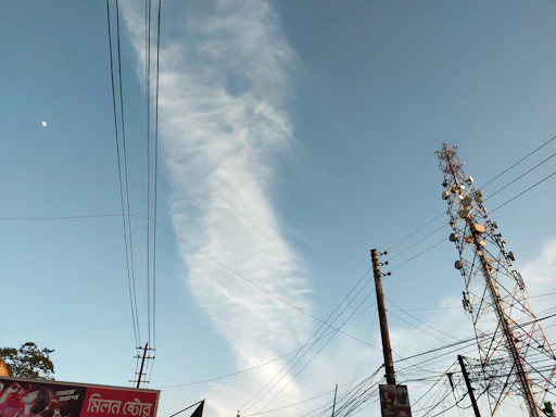 A tall telecommunications tower stands against a clear blue sky with a wisp of white cloud. Several power lines are visible, crisscrossing the scene. Part of an advertising billboard is visible in the corner with text in a foreign script.