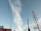 A tall telecommunications tower stands against a clear blue sky with a wisp of white cloud. Several power lines are visible, crisscrossing the scene. Part of an advertising billboard is visible in the corner with text in a foreign script.
