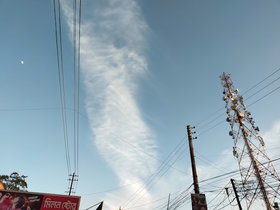 A tall telecommunications tower stands against a clear blue sky with a wisp of white cloud. Several power lines are visible, crisscrossing the scene. Part of an advertising billboard is visible in the corner with text in a foreign script.