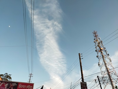 A tall telecommunications tower stands against a clear blue sky with a wisp of white cloud. Several power lines are visible, crisscrossing the scene. Part of an advertising billboard is visible in the corner with text in a foreign script.