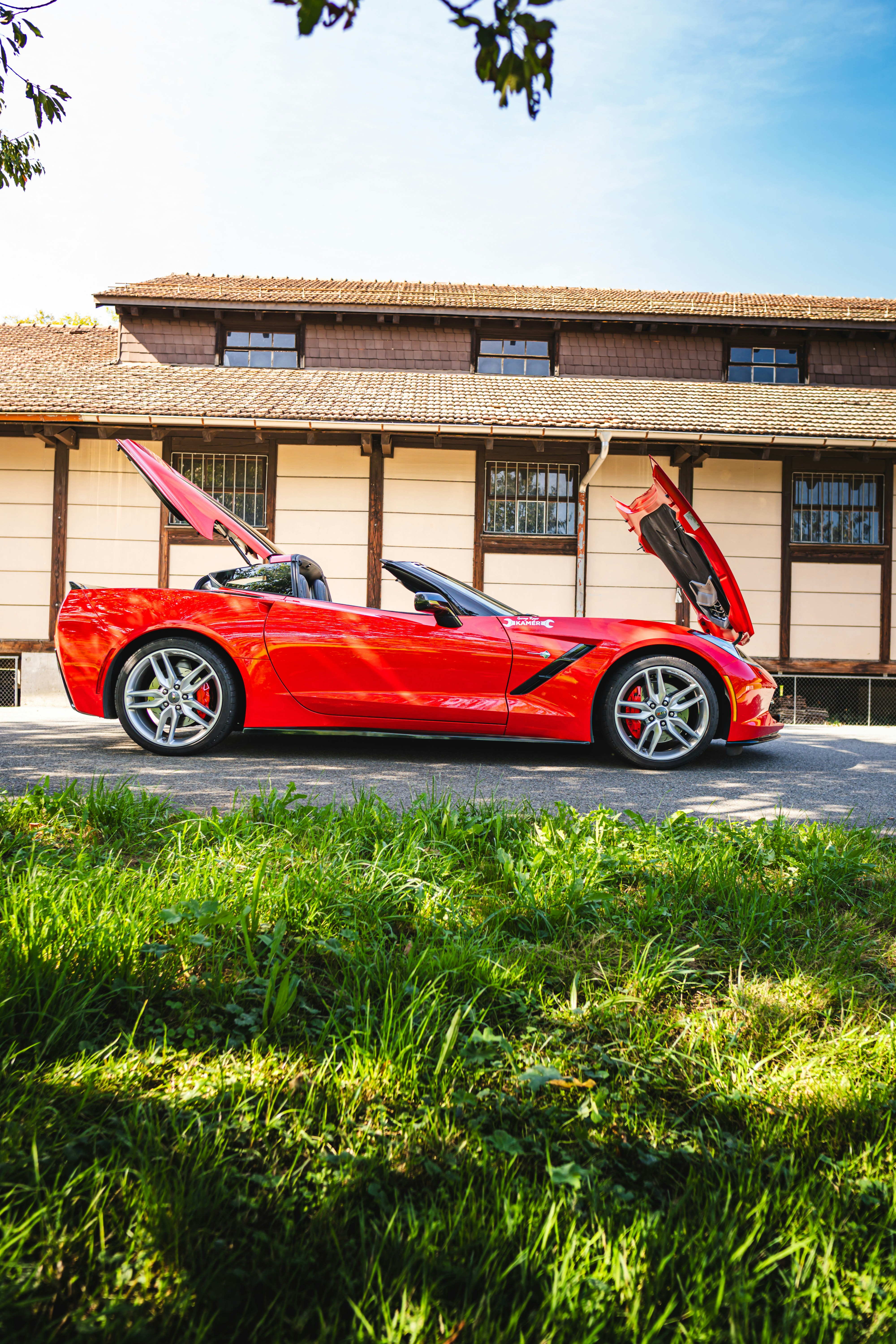 A red convertible sports car with its hood and trunk raised sits in front of a timber-framed building, under a bright blue sky. Lush grass in the foreground adds depth.