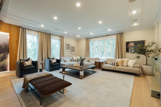 A spacious and modern living room with a blend of neutral tones. The room features a large beige sectional sofa adorned with pillows, a dark brown bench, and two black armchairs. A central coffee table displays decorative items, including a small plant. The room is illuminated by recessed lighting and natural light from three large windows with beige curtains. Artwork and a potted plant add a sophisticated touch to the decor.