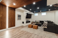 A cozy living room features a large sectional sofa adorned with cushions and throws, positioned in a well-lit space with a wooden accent wall. The room includes a modern fireplace and a large wall-mounted television. The flooring is light wood, and a geometric patterned rug adds texture. Two brown leather ottomans serve as additional seating or footrests.