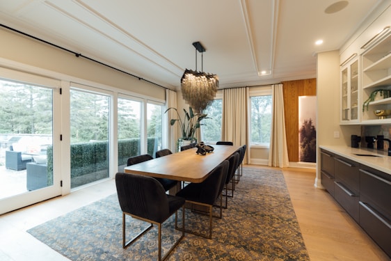 A modern dining room with a long rectangular wooden table surrounded by black upholstered chairs. The room features large sliding glass doors on the left, providing ample natural light and views of greenery outside. A decorative chandelier hangs above the table, and a patterned rug covers the wooden floor. On the right, a sleek kitchen counter with cabinets and open shelves displays a few decorative items and a potted plant.