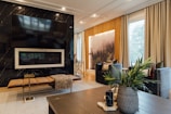 A modern living room featuring a large flat-screen TV mounted on a black marble wall above a sleek fireplace. A long cushioned bench sits in front with a textured throw draped over it. The room is warmly lit and includes comfortable dark blue armchairs with patterned pillows, a wooden coffee table with decorative items, and lush green plants in a vase. A backlit forest landscape art piece adds a serene touch to the room, positioned between large windows covered with beige curtains.