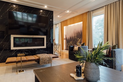 A modern living room featuring a large flat-screen TV mounted on a black marble wall above a sleek fireplace. A long cushioned bench sits in front with a textured throw draped over it. The room is warmly lit and includes comfortable dark blue armchairs with patterned pillows, a wooden coffee table with decorative items, and lush green plants in a vase. A backlit forest landscape art piece adds a serene touch to the room, positioned between large windows covered with beige curtains.
