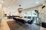 A modern dining area seamlessly connected to a kitchen, featuring a long wooden table surrounded by dark upholstered chairs. A large, unique chandelier hangs above the table, providing elegant lighting. The space is filled with natural light from a wall of large windows. Indoor plants add a touch of greenery, while a patterned rug under the dining table adds texture. The kitchen area in the background is equipped with sleek appliances and additional dining seating at an island.
