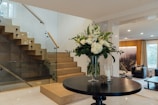 A modern, stylish interior with a wooden staircase featuring glass railings and a small round table with a vase of white and green flowers. The room has a warm ambiance with soft lighting and a natural landscape artwork on the wall. Comfortable seating and large windows add to the cozy atmosphere.