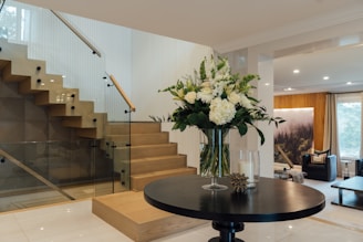 A modern, stylish interior with a wooden staircase featuring glass railings and a small round table with a vase of white and green flowers. The room has a warm ambiance with soft lighting and a natural landscape artwork on the wall. Comfortable seating and large windows add to the cozy atmosphere.