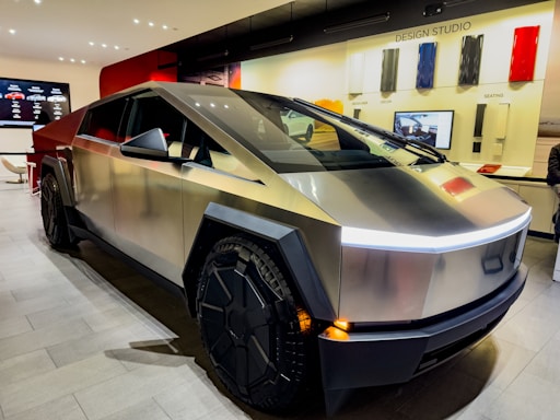 A futuristic, angular vehicle with a metallic finish is parked indoors in a modern showroom. The vehicle features large, distinctive wheels, and the showroom has a minimalist design with a display screen and color samples on the walls.