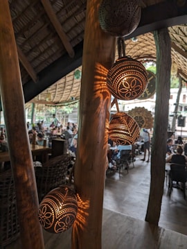 A cozy, rustic indoor space with wicker furniture and a roof made from natural materials. There is a wooden support structure visible in the foreground, adorned with decorative spherical lamps that emit a warm, soft light. The background includes seated people in a communal dining setting, giving the atmosphere a relaxed and social vibe.