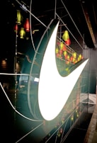 A large, illuminated swoosh logo is prominently displayed against a dark, reflective wall, with multiple colorful lanterns in shades of red and green hanging and reflecting across the surface.