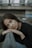 A woman with long brown hair leans her head on her folded hands against a railing. Her expression is introspective and calm, and she is dressed in a black top with a blurred background of an urban setting.