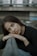 A woman with long brown hair leans her head on her folded hands against a railing. Her expression is introspective and calm, and she is dressed in a black top with a blurred background of an urban setting.