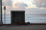 A weathered, empty beach bunker or shelter overlooks the ocean waves crashing against the shore. A worn-out armchair is placed outside the structure, and there is a garbage bin nearby. The sky is overcast with clouds that reflect soft shades of orange and pink from the setting or rising sun. A sign and power pole are visible to the left.