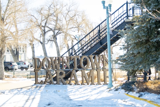 A snowy scene featuring a large metal sign that reads 'Downtown Laramie' in front of a black staircase. Leafless trees and a few parked cars are visible in the background, along with a green pine tree and a streetlamp in the foreground.