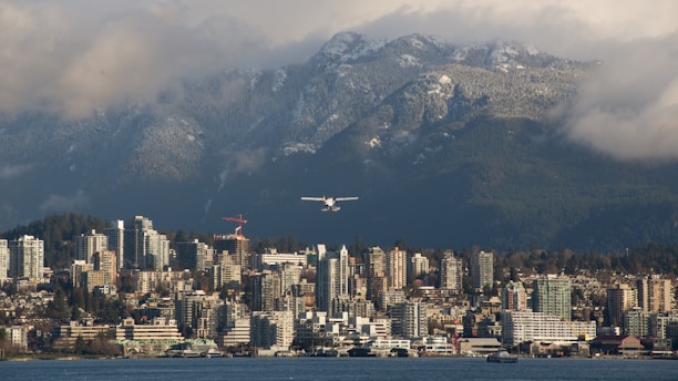 A city skyline with a backdrop of majestic mountains partially covered in snow. A seaplane is flying above the cityscape, and some clouds hang in the sky.