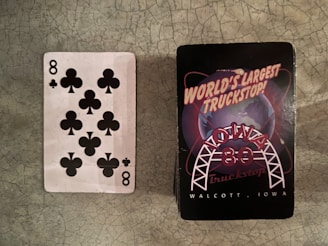 A playing card showing the 8 of clubs is placed next to a deck of cards with a cover that reads 'World's Largest Truckstop, Iowa 80, Walcott, Iowa'. The background surface appears to be a textured, cracked material.