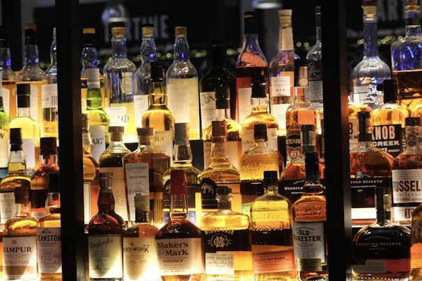 A collection of various whiskey bottles is displayed on shelves, illuminated warmly from behind. The bottles vary in shape, size, and label design, showcasing different brands and types. The lighting creates a cozy and inviting ambiance.