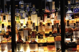 A collection of various whiskey bottles is displayed on shelves, illuminated warmly from behind. The bottles vary in shape, size, and label design, showcasing different brands and types. The lighting creates a cozy and inviting ambiance.