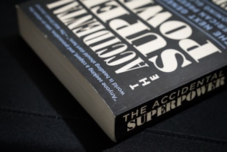 A book titled 'The Accidental Superpower' lies at an angle on a dark surface. The cover features large, bold white text on a black background, with additional smaller text running along the spine of the book.