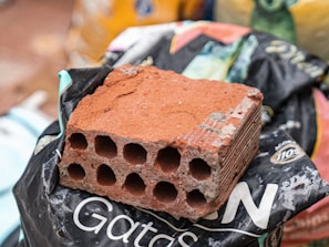 A red, rectangular brick with multiple circular holes is placed on a black plastic bag. The black bag has some white letters and logos. There are various bags and objects blurred in the background.