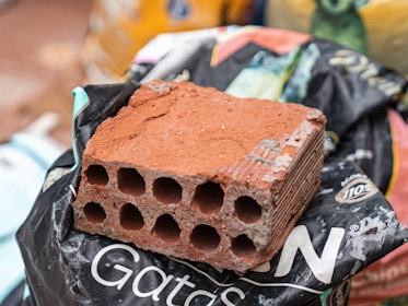 A red, rectangular brick with multiple circular holes is placed on a black plastic bag. The black bag has some white letters and logos. There are various bags and objects blurred in the background.