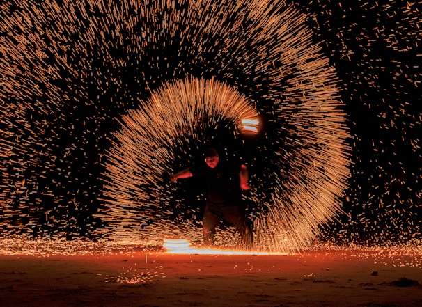 A person is performing a fire dance at night, creating mesmerizing arcs of glowing sparks that illuminate the darkness. The fiery trails form a circular pattern around the performer, who is dynamically caught in action with blurred motion.