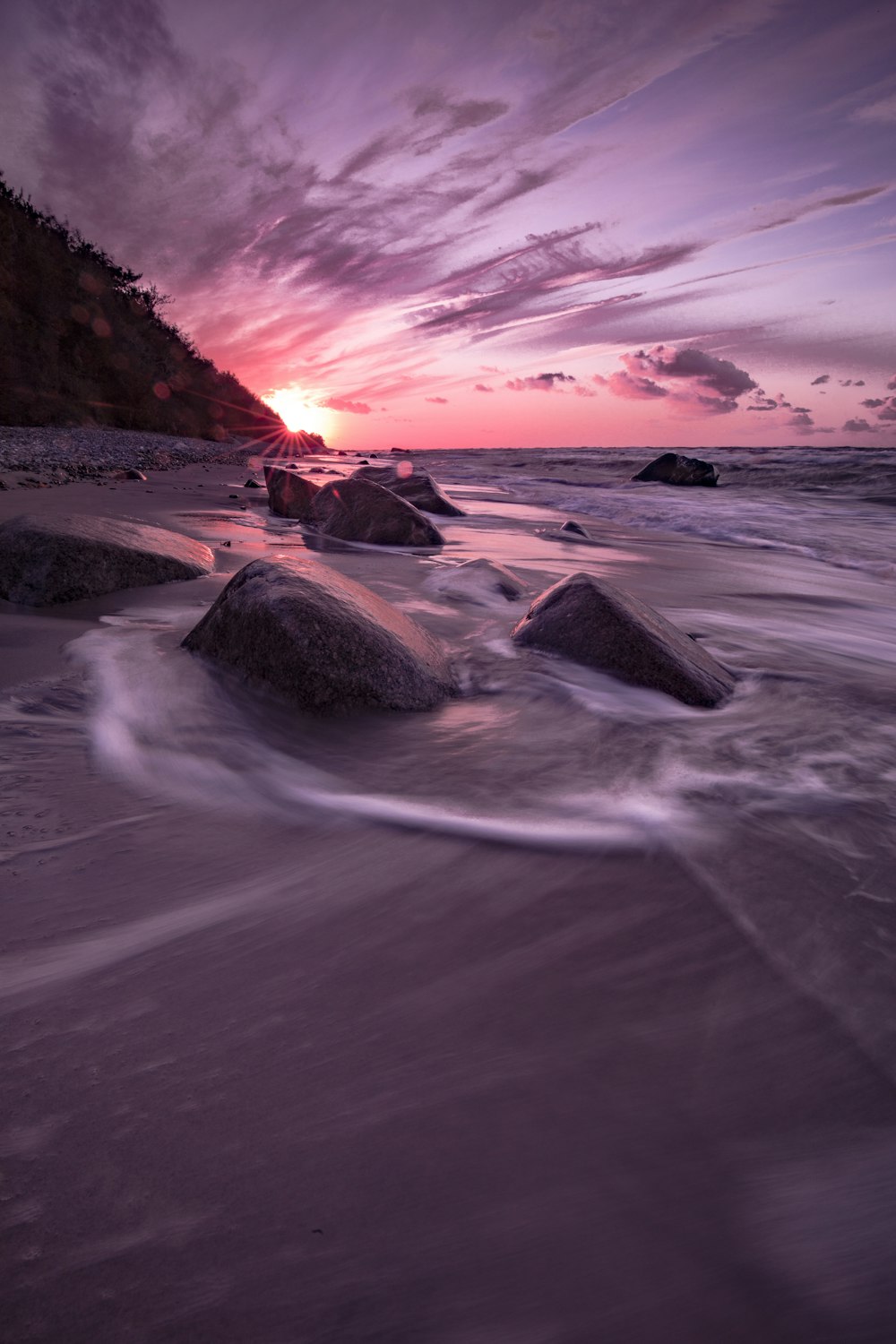 the sun is setting over the ocean with rocks in the foreground