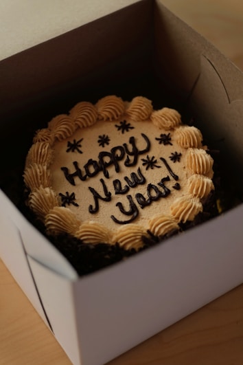 A round cake inside a box, decorated with light brown frosting along the edges and the message 'Happy New Year!' written in dark icing on top.