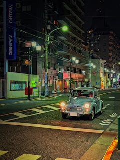A vintage car drives down an urban street at night. The scene is illuminated by streetlights and neon signs from surrounding buildings. There is a mix of traditional and modern architectural elements, and a bank sign is visible.