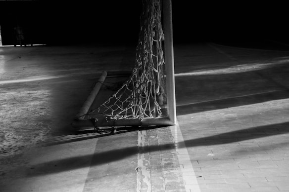 A black and white image of a sports goal with a net attached, casting dramatic shadows on a hard floor surface. The netting creates interesting geometric patterns with the light and shadows.