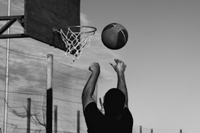 A monochrome image of a person shooting a basketball towards the hoop with the ball captured mid-air. The basketball hoop is visible against a clear sky, and there is a wire fence in the background.
