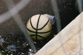 A basketball lies on a gritty, littered surface next to a plastic cup, surrounded by small debris and hints of nature.