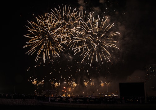 Bright and large fireworks burst in the night sky, creating intricate patterns with golden sparks. The scene is lively, and there is a dark, silhouetted foreground that includes spectators viewing the display.
