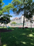 A lush green park area with large trees providing shade. Colorful benches are scattered on the grass, and a swing set is visible on the left side. In the background, a row of historic buildings with red and white facades line the street. Bicycles are parked along the sidewalk, and a clear blue sky is above.