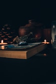 A dimly lit scene featuring a thick book lying on a surface, surrounded by small lit candles. There are herbs resting on the book and a large pine cone alongside. In the background, a rustic clay pot is visible, adding to the warm and cozy ambiance.