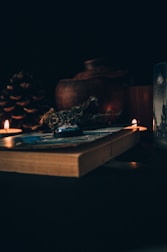 A dimly lit scene featuring a thick book lying on a surface, surrounded by small lit candles. There are herbs resting on the book and a large pine cone alongside. In the background, a rustic clay pot is visible, adding to the warm and cozy ambiance.