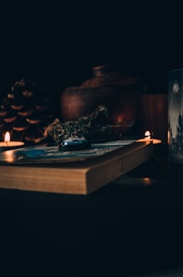 A dimly lit scene featuring a thick book lying on a surface, surrounded by small lit candles. There are herbs resting on the book and a large pine cone alongside. In the background, a rustic clay pot is visible, adding to the warm and cozy ambiance.