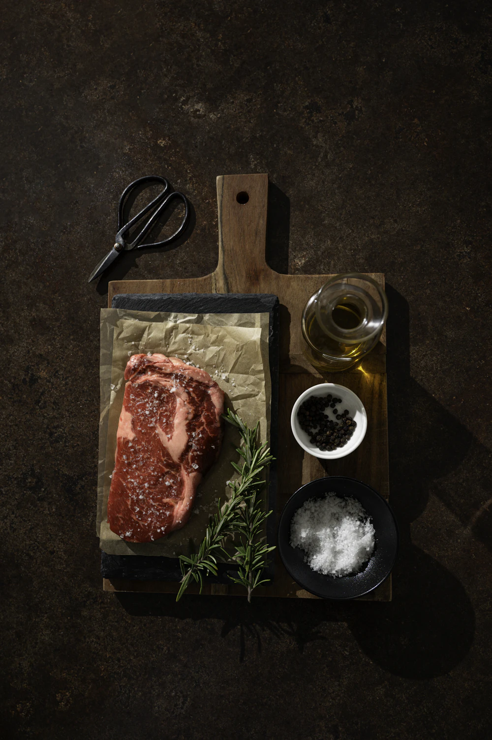 Bife crudo sobre papel en una tabla, con romero, sal gruesa y pimienta