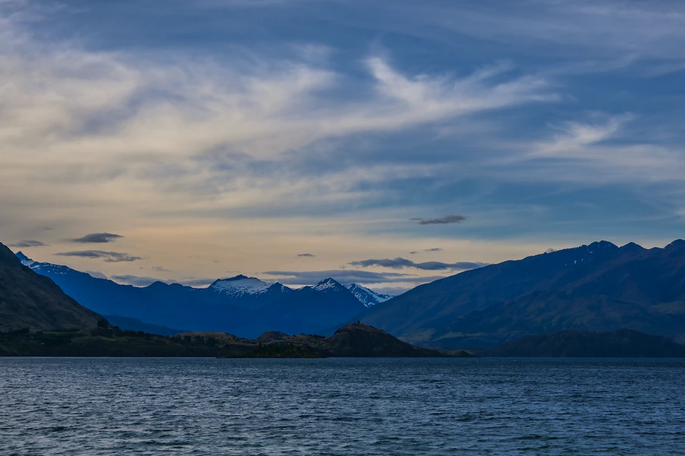 The image depicts a serene mountain range with snow-capped peaks and deep blue water below, creating a striking contrast between the natural elements. The mountains are bathed in hues of blue and white, while the water mirrors these colors, adding to the overall beauty of the scene. The perspective is from across the body of water, providing a panoramic view of the majestic landscape.